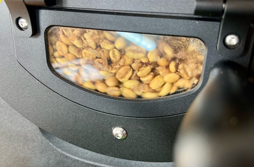 Grüne Kaffeebohnen drehen sich in der Trommel eines professionellen Kaffeerösters. Durch das Sichtfenster erkennt man die erste Phase des Röstprozesses – die Bohnen beginnen langsam, ihre Farbe zu verändern und erste Röstaromen zu entwickeln. Die Szene vermittelt den handwerklichen Charakter der Kaffeeveredelung., © ertl.kaffee